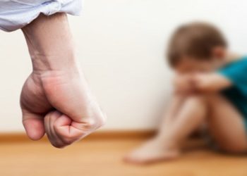 Family violence and aggression concept - furious angry man raised punishment fist over scared or terrified child boy sitting at wall corner